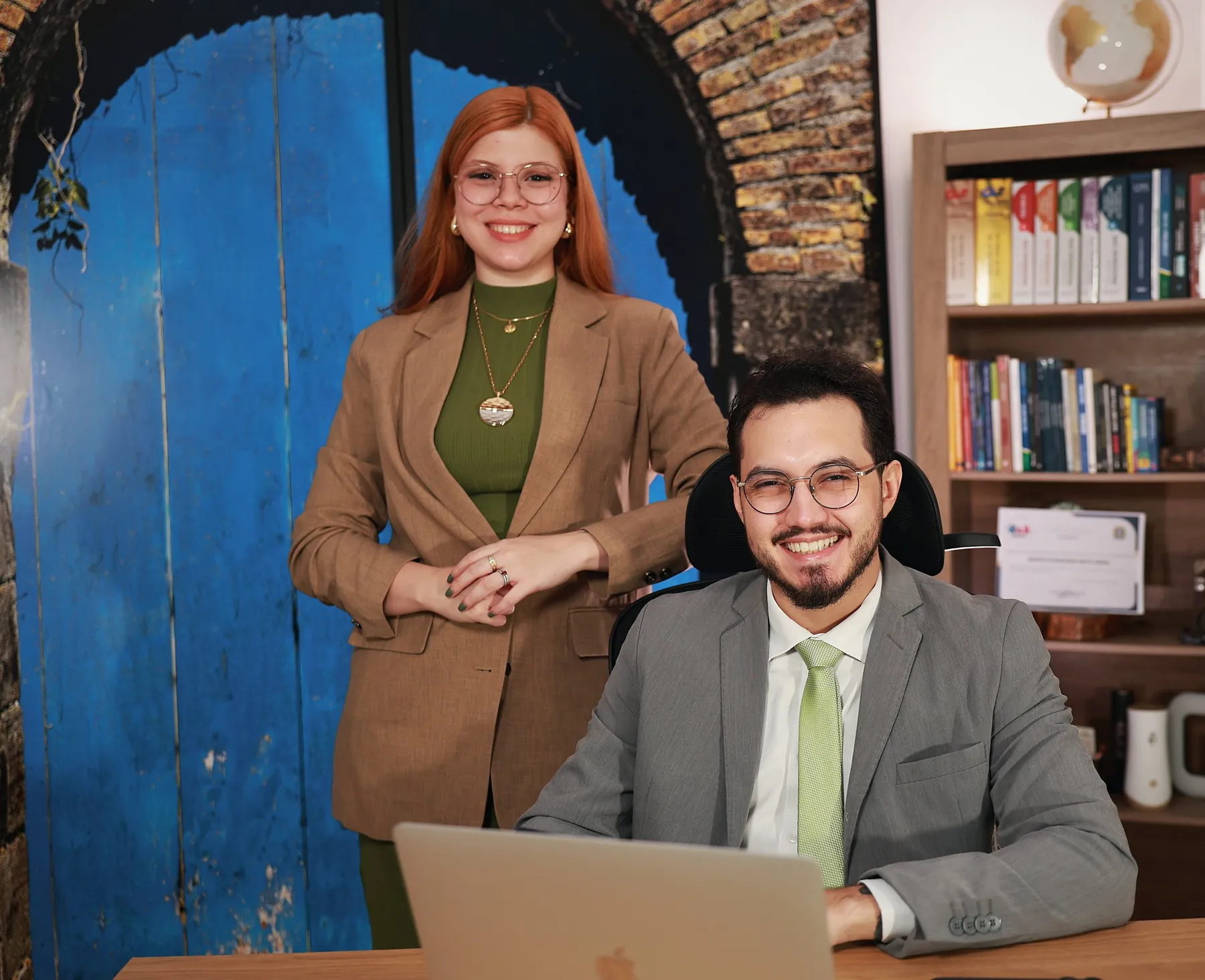 Rita Montenegro, em pé, e Mateus Serra, sentado à mesa, sorrindo no escritório